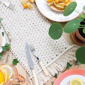 Peut inclure: Un chemin de table en crochet blanc avec des franges sur les bords. Le chemin de table est pos&eacute; sur une table en bois avec une fourchette et un couteau dessus. Il y a des plantes et des fruits en arri&egrave;re-plan.
