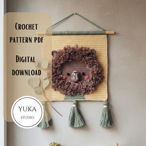 May include: A crochet lion wall hanging with a textured brown mane, pink cheeks, and a detailed face. The wall hanging is yellow and has green tassels at the bottom. The text "CROCHET PATTERN PDF DIGITAL DOWNLOAD" is visible.