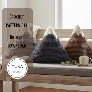 May include: Three crochet mountain-shaped pillows in gray, navy, and brown with white peaks, arranged on a wooden bench. A mug of coffee sits nearby. The image includes text that reads "CROCHET PATTERN PDF DIGITAL DOWNLOAD" and "YUKA STUDIO."