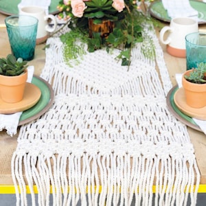 K&ouml;nnte beinhalten: Ein wei&szlig;er geh&auml;kelter Tischl&auml;ufer mit Fransen auf einem Holztisch. Der L&auml;ufer hat ein Rautenmuster und ist mit Gr&uuml;npflanzen und Blumen verziert.