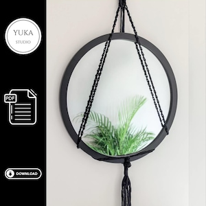 Può includere: Uno specchio rotondo nero con un design a sospensione in macramè. Lo specchio riflette una pianta verde. È sospeso da una corda nera e ha un nappa decorativa. Il testo "YUKA STUDIO" è in un cerchio bianco in alto a sinistra.
