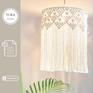 Peut inclure: Un abat-jour en macramé blanc avec une base à franges et un motif géométrique sur le dessus. Le luminaire est suspendu au plafond et dégage une lueur douce et chaleureuse. Le texte "YUKA STUDIO" est dans un cercle à gauche.