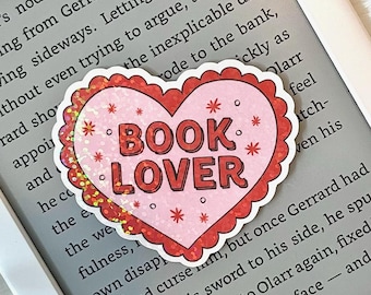 May include: A pink heart-shaped sticker with glitter and the words "BOOK LOVER" in red letters. The sticker is placed on a gray background with text.