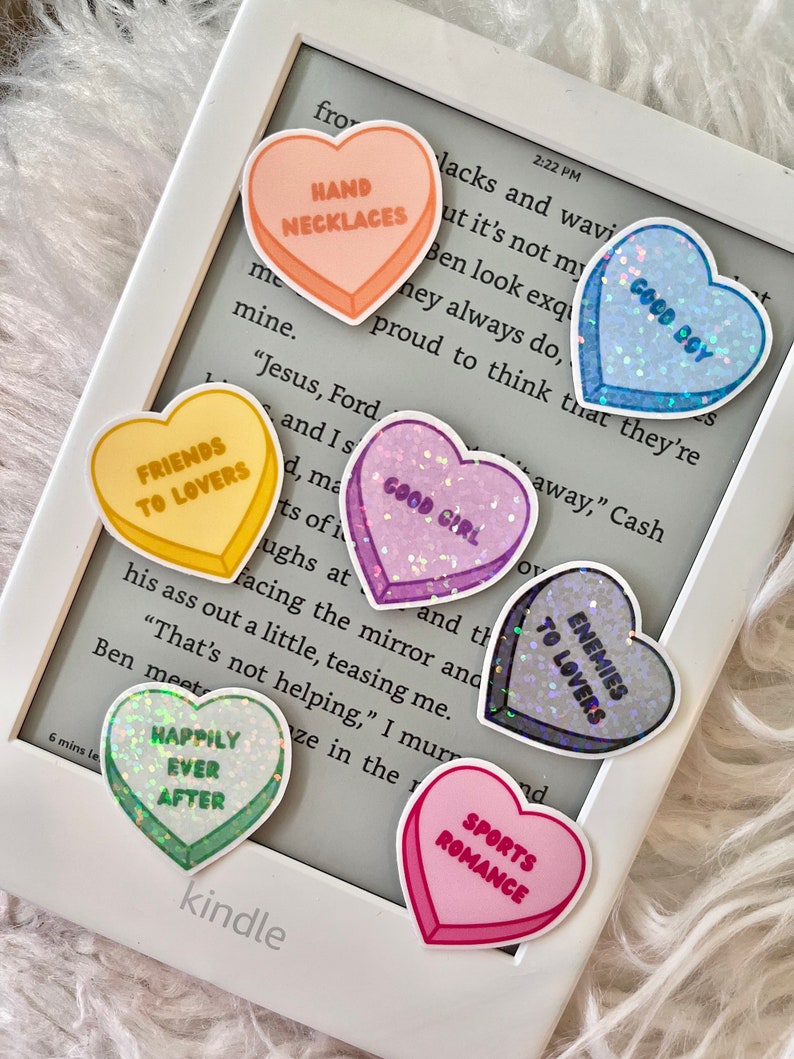 May include: A collection of heart-shaped stickers with various sayings, including "Hand Necklaces", "Good Boy", "Good Girl", "Friends to Lovers", "Enemies to Lovers", "Happily Ever After", and "Sports Romance". The stickers are on a white Kindle e-reader.