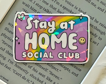May include: A holographic sticker with the text "Stay at HOME SOCIAL CLUB" in white, bubble letters on a purple background. A rainbow design is above the word "HOME", and a yellow smiley face is on the right.