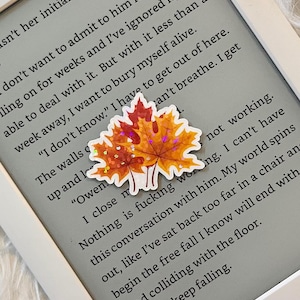 May include: A close-up of a Kindle e-reader displaying a page of text with a fall-themed sticker of two orange and red leaves with glitter accents. The text reads "I close my nothing is fucking working. I can't have this conversation with him. My world spins out, like I've sat back too far in a chair and begin the free fall I know will end with head colliding with the floor. But I keep falling."