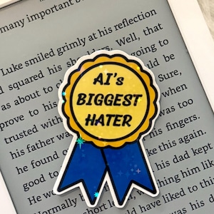 May include: A sticker shaped like a circular award ribbon with a yellow centre and blue tails. The yellow centre features the black text AI'S BIGGEST HATER. The sticker has a holographic finish with small star accents, placed on a white e-reader screen.