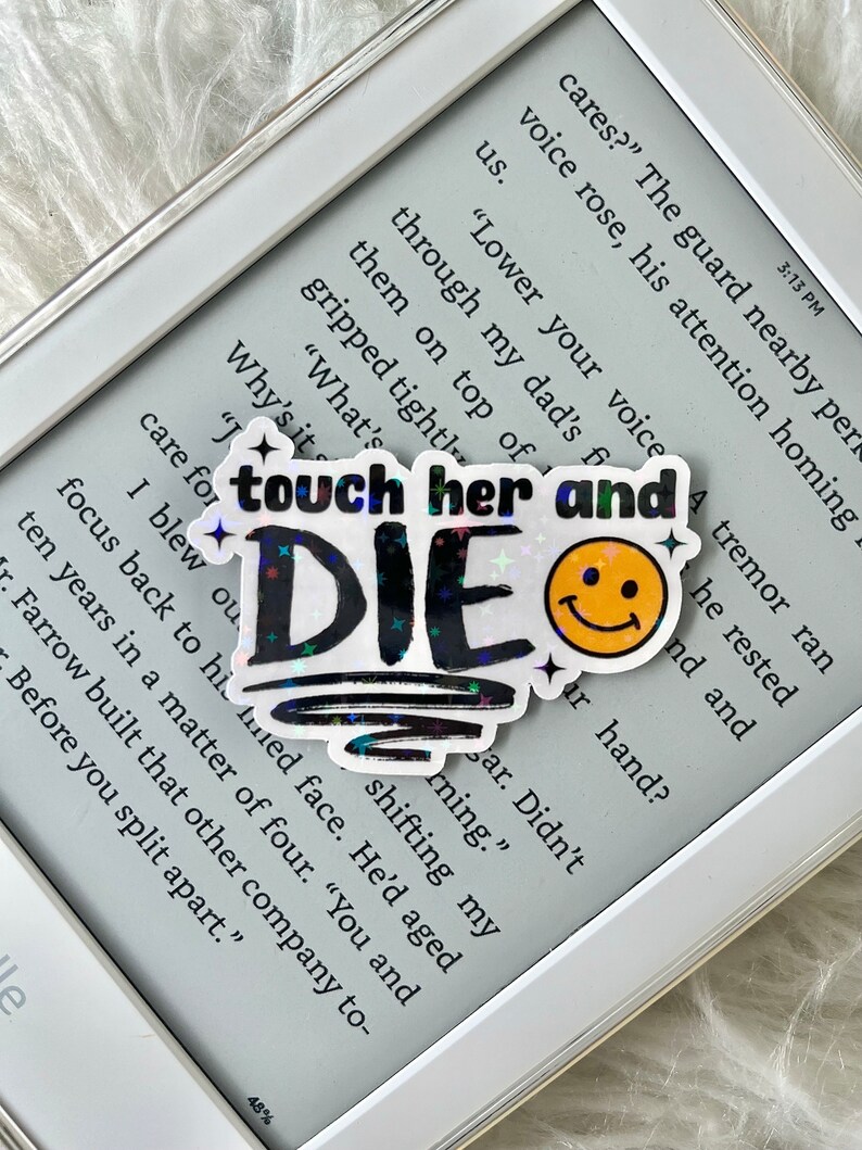 May include: A holographic sticker with the text "touch her and die" in black with rainbow glitter. A yellow smiley face sticker is next to it. The sticker is on a white e-reader.