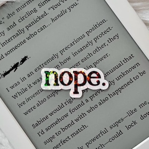 May include: A close-up of a rainbow glitter sticker with the word "nope" in black letters. The sticker is placed on a page of a book with text about a precarious position.