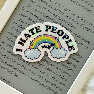 Puede incluir: Una pegatina blanca con el texto "I HATE PEOPLE" en negro, contorneada en plata. La pegatina presenta un arcoíris, nubes y un murciélago. La pegatina está colocada en un lector Kindle.
