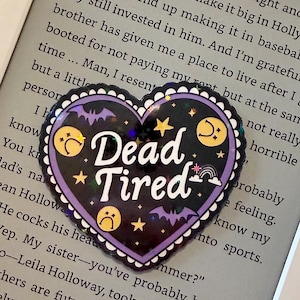 May include: A heart-shaped sticker with the words "Dead Tired" in white. The sticker has a black background with purple accents, bats, stars, moons, and sad face emojis. The sticker is on a gray surface with text.