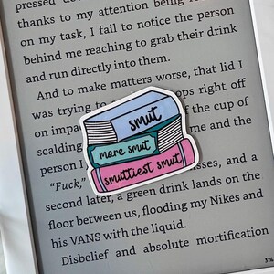 May include: A sticker of a stack of books with the words "smut", "more smut", and "smuttiest smut" on them. The sticker is placed on a Kindle e-reader with the text "attempting to fix the lid that's not pressed down completely. Of course, thanks to my attention being focused on my task, I fail to notice the person behind me reaching to grab their drink and run directly into them. And to make matters worse, that lid I was trying to... ops right off the cup of... me and the... "Fuck," ... second later, a green drink lands on the floor between us, flooding my Nikes and his VANS with the liquid. Disbelief and absolute mortification"