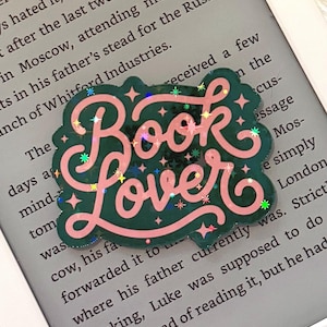 May include: A dark green acrylic pin with the text Book Lover in pink cursive lettering. The pin features a holographic finish with small star-shaped accents scattered across the surface, resting on the screen of an e-reader displaying text.