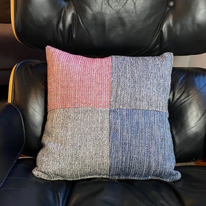 May include: A square throw pillow with a patchwork design. The pillow is made of a textured fabric and has four panels in different colors: pink, gray, blue, and gray. The pillow is sitting on a black leather chair.