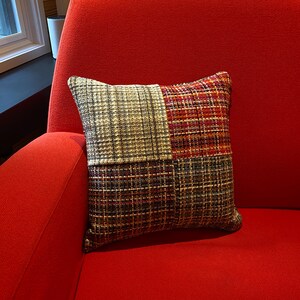Puede incluir: Cojín decorativo cuadrado con diseño de patchwork. El cojín presenta cuatro secciones con diferentes patrones de cuadros en tonos marrón, rojo y beige. El cojín está sobre una silla roja.