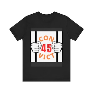 Puede incluir: Camiseta negra con un diseño gráfico que presenta la palabra "CONVICT" con el número "45" en el centro. El diseño incluye un par de manos de dibujos animados agarrando barrotes de prisión.