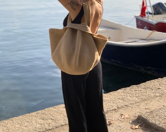 May include: A woman wearing black pants and a black swimsuit is standing on a dock by the water. She is carrying a large, beige crocheted tote bag.