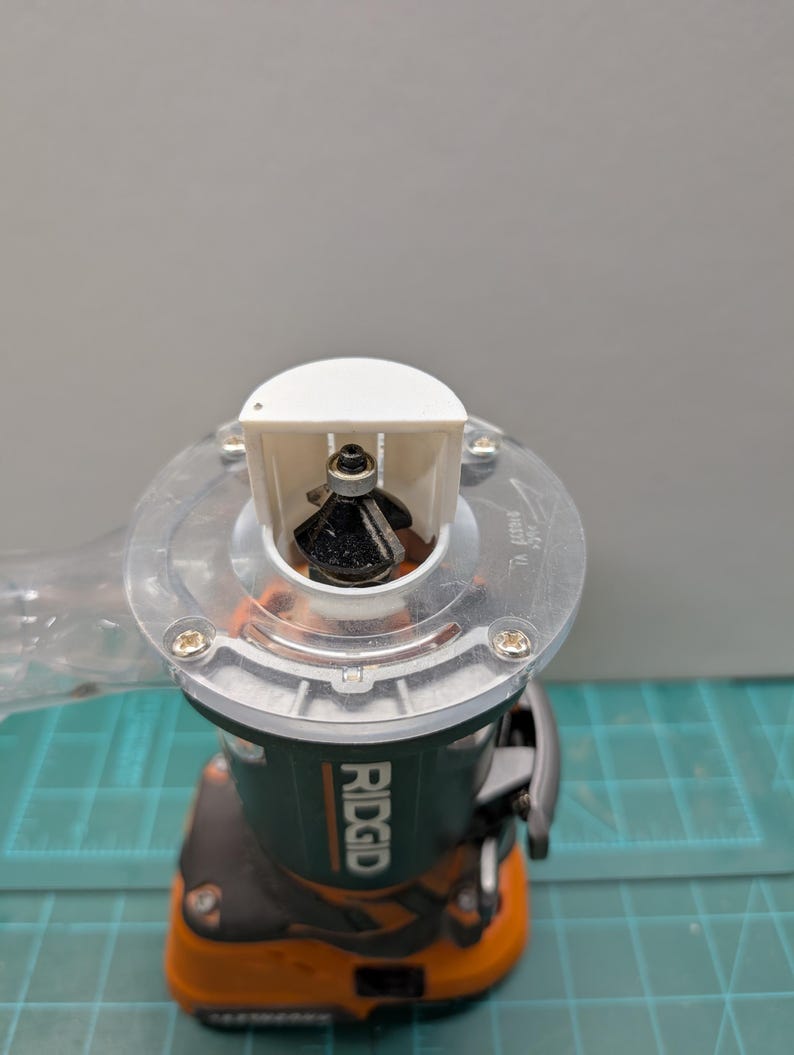 Pu&ograve; includere: Primo piano di una fresatrice del marchio Ridgid. La fresatrice ha una base in plastica trasparente, un corpo nero e arancione e una protezione bianca. La punta della fresatrice &egrave; nera e visibile all'interno della protezione.