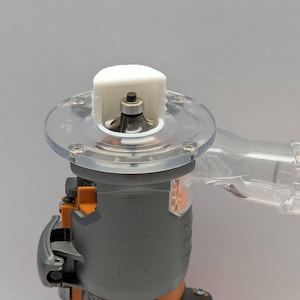 Pu&ograve; includere: Un elettroutensile grigio e arancione con una base in plastica trasparente e una parte superiore bianca. L'utensile ha una porta di raccolta della polvere in plastica trasparente. Progettato per la lavorazione del legno e altri progetti fai-da-te.