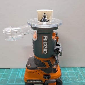 Pu&ograve; includere: Una fresatrice a batteria RIDGID arancione e nera con un accessorio di raccolta della polvere in plastica trasparente. La fresatrice ha una punta argentata e un'impugnatura nera.
