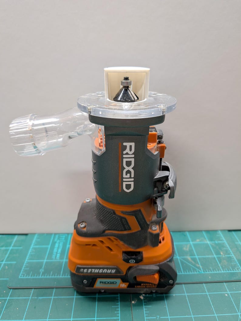 Pu&ograve; includere: Una fresatrice a batteria RIDGID arancione e nera con un accessorio di raccolta della polvere in plastica trasparente. La fresatrice ha una punta argentata e un'impugnatura nera.