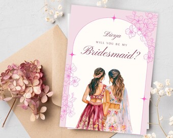 May include: A pink bridesmaid proposal card with the text "Divya, Will You Be My Bridesmaid?" features illustrations of two women in traditional Indian attire. The card is surrounded by floral accents and rests on a neutral surface.