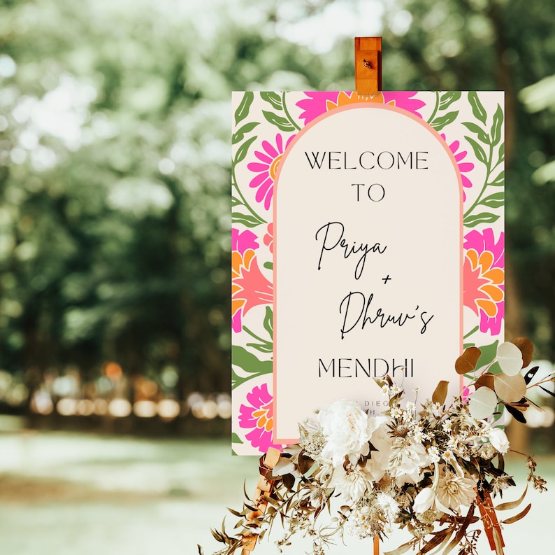 May include: A welcome sign for a Mehndi celebration with a floral design in pink, orange, and green. The sign reads "Welcome to Priya + Dhruv's Mehndi".