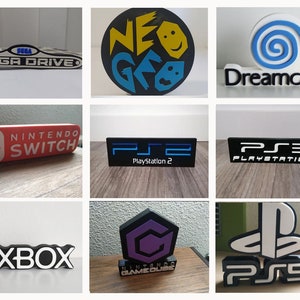 Puede incluir: Una colección de logotipos de consolas de videojuegos, incluyendo Sega Mega Drive, Neo Geo, Dreamcast, Nintendo Switch, PlayStation 2, PlayStation 3, Xbox, Nintendo GameCube y PlayStation 5. Cada logotipo es un diseño impreso en 3D.