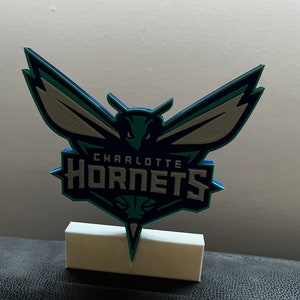 Puede incluir: Un logotipo de los Charlotte Hornets impreso en 3D con un esquema de color azul verdoso, azul y blanco. El logotipo presenta un avispón estilizado con las alas extendidas y el texto "Charlotte Hornets" debajo.