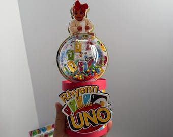 Könnte beinhalten: Ein personalisiertes Party-Tischgesteck mit einer transparenten Kunststoffkuppel, gefüllt mit bunten Perlen und Kartenmotiven. Obenauf befindet sich ein Kinderausschnitt mit roter Schleife. Der rosa Sockel trägt die Aufschrift Rhyenn turns UNO.