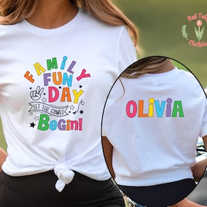May include: White t-shirt with the text "FAMILY FUN DAY" in colorful letters and the phrase "LET THE GAMES BEGIN!". The back of the shirt has the name "OLIVIA" in colorful letters. The shirt is tied at the waist.