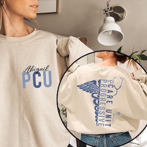 May include: Cream-colored sweatshirt with the name "Abigail" and "PCU" in blue lettering on the front. The back features a blue caduceus design with the words "PCU Care Unit Progressive" and a stethoscope.