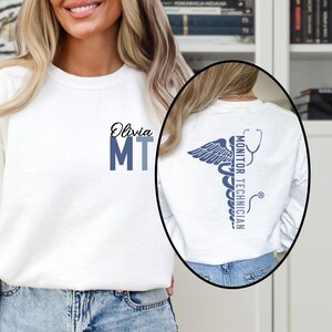 May include: White sweatshirt with the name "Olivia" and "MT" in blue on the front. The back features a blue caduceus design with the words "Monitor Technician".