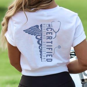 以下が含まれることがあります： 青いカドゥケウスのシンボルと「CNA CERTIFIED NURSING ASSISTANT」の文字が入った白いTシャツ。デザインには聴診器が含まれています。シャツは柔らかい素材で作られています。