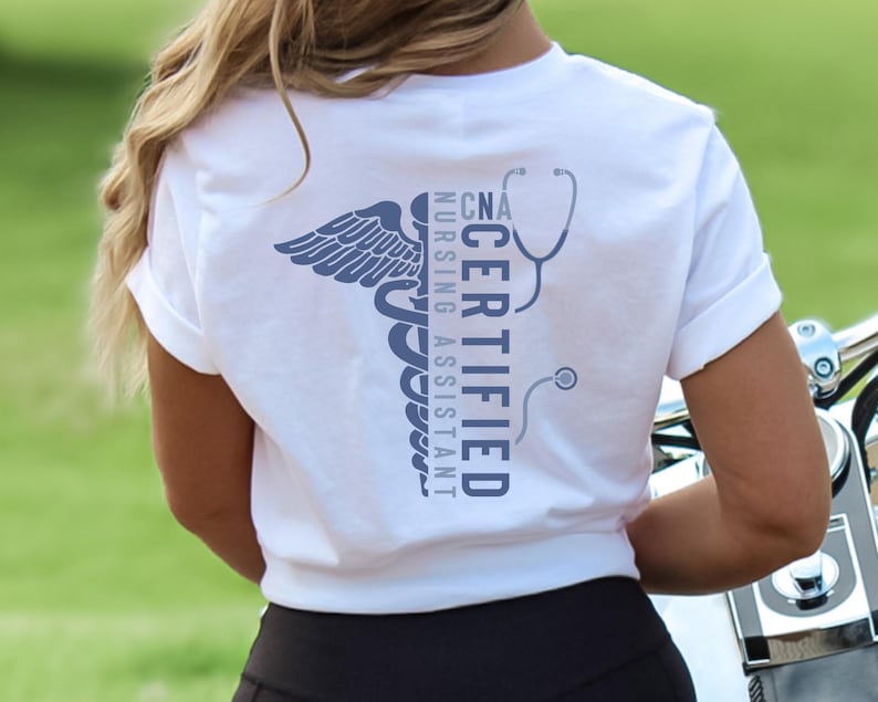 以下が含まれることがあります： 青いカドゥケウスのシンボルと「CNA CERTIFIED NURSING ASSISTANT」の文字が入った白いTシャツ。デザインには聴診器が含まれています。シャツは柔らかい素材で作られています。