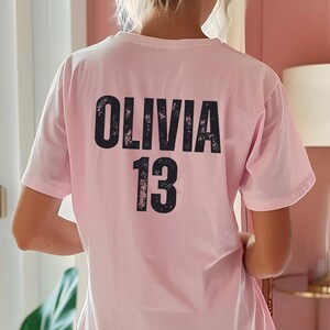 May include: Pink t-shirt with black text that says "Olivia 13" on the back.