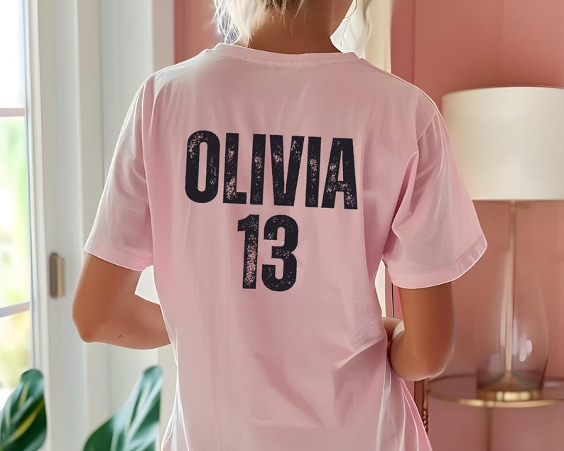 May include: Pink t-shirt with black text that says "Olivia 13" on the back.