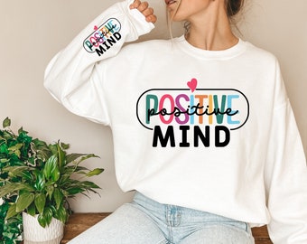 May include: White sweatshirt with a colorful "POSITIVE MIND" graphic on the front. The graphic features a pink heart and a rainbow-colored word "positive" inside a black oval.
