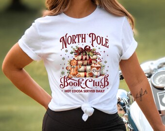 May include: White t-shirt featuring a festive design for a North Pole Book Club. The graphic includes stacked books, a gingerbread man, and a mug of cocoa. Text reads "North Pole Book Club" and "Hot Cocoa Served Daily".