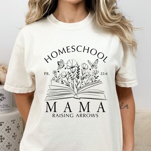 Puede incluir: Camiseta color crema con un gr&aacute;fico negro. El gr&aacute;fico presenta las palabras "HOMESCHOOL MAMA RAISING ARROWS" sobre un libro abierto con flores y mariposas. Tambi&eacute;n se incluye el texto "PR. 22:6".