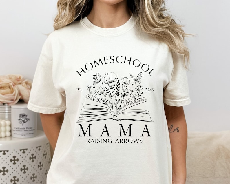 Puede incluir: Camiseta color crema con un gr&aacute;fico negro. El gr&aacute;fico presenta las palabras "HOMESCHOOL MAMA RAISING ARROWS" sobre un libro abierto con flores y mariposas. Tambi&eacute;n se incluye el texto "PR. 22:6".