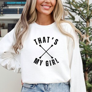 May include: White sweatshirt with the text "THAT'S MY GIRL" in black, with crossed batons. The sweatshirt is long-sleeved and has a crew neck. The design is centered on the front of the shirt.