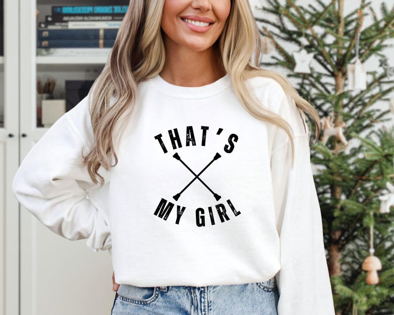 May include: White sweatshirt with the text "THAT'S MY GIRL" in black, with crossed batons. The sweatshirt is long-sleeved and has a crew neck. The design is centered on the front of the shirt.