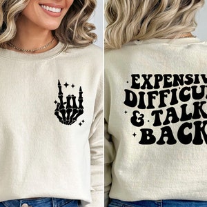 K&ouml;nnte beinhalten: Ein cremefarbener Pullover mit einer schwarzen Skeletthand, die ein Rock'n'Roll-Zeichen auf der Vorderseite macht, und dem Text "EXPENSIVE DIFFICULT & TALKS BACK" auf der R&uuml;ckseite.