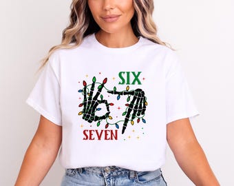 May include: White t-shirt featuring a skeleton hand design with Christmas lights. The words "SIX" and "SEVEN" are printed in green and red, respectively. The design incorporates colorful lights, creating a festive and spooky aesthetic.