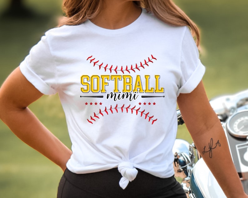May include: White t-shirt with a red and yellow graphic that says "Softball mimi" with a baseball bat and stitching around the text.