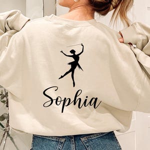 May include: Beige sweatshirt with a black silhouette of a baton twirler and the name "Sophia" in cursive script. The sweatshirt has long sleeves and a crew neck. The twirler is holding a baton above their head.