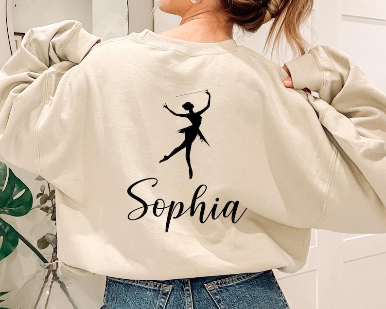 May include: Beige sweatshirt with a black silhouette of a baton twirler and the name "Sophia" in cursive script. The sweatshirt has long sleeves and a crew neck. The twirler is holding a baton above their head.