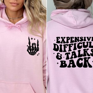 K&ouml;nnte beinhalten: Rosafarbener Hoodie mit einer schwarzen Skeletthand, die ein Rock'n'Roll-Zeichen auf der Vorderseite macht, und dem Text "EXPENSIVE DIFFICULT & TALKS BACK" auf der R&uuml;ckseite.