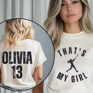 May include: Two cream colored t-shirts with black text. One shirt says "Olivia 13" and the other says "That's My Girl" with a silhouette of a girl playing baseball.